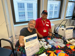Two GMHC staff members with a table of resources on mental health and insurance access.